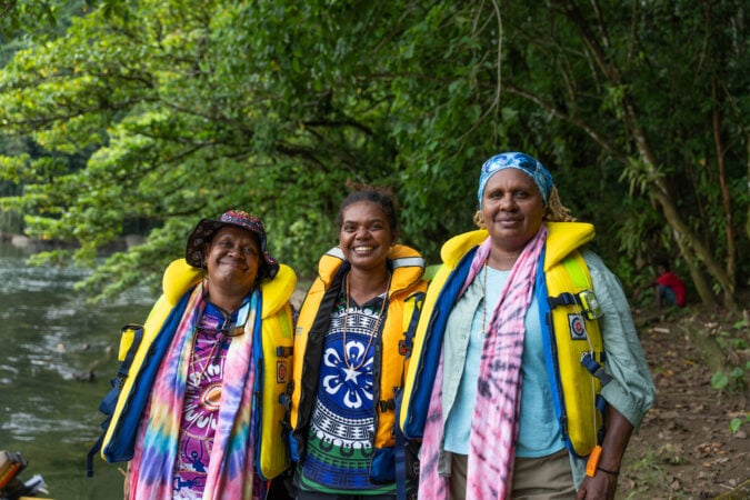 Three proud Indigenous women in the Solomon Islands including Torres Strait Islander rangers wearing yellow life vests with trees and water in background