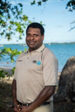 A smiling Torres Strait Islander man in ranger uniform infront of ocean of the Torres Strait region.