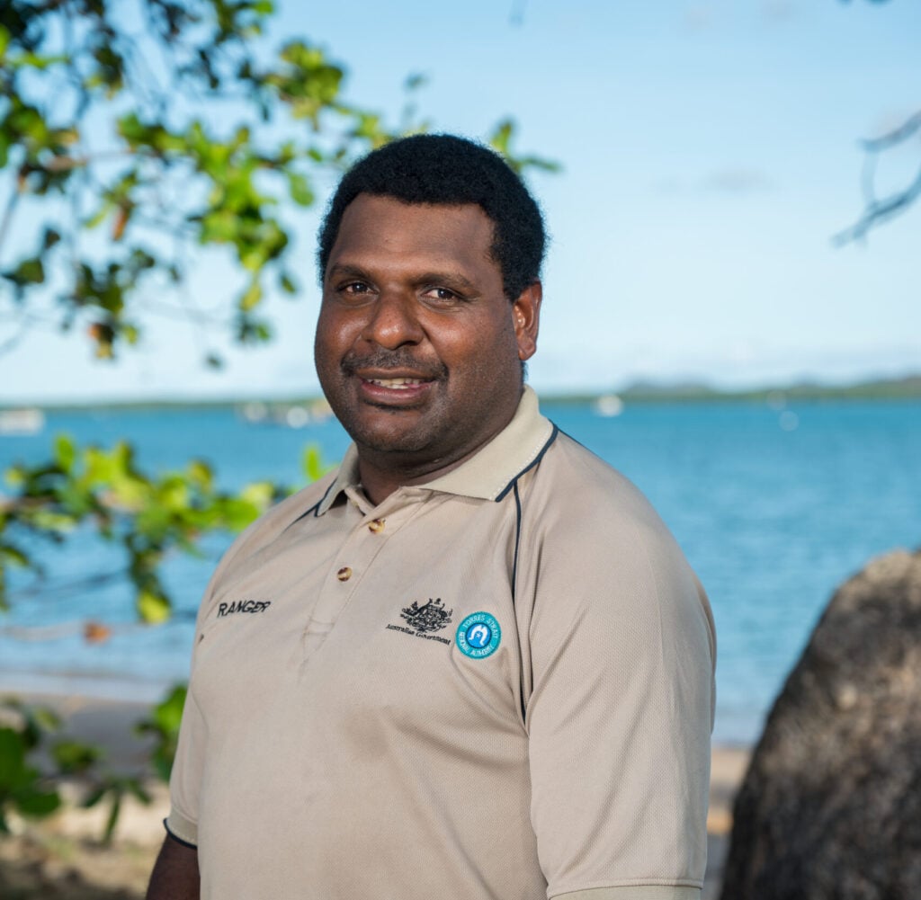 A smiling Torres Strait Islander man in ranger uniform infront of ocean of the Torres Strait region.