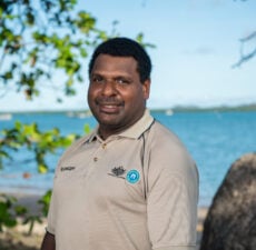 A smiling Torres Strait Islander man in ranger uniform infront of ocean of the Torres Strait region.